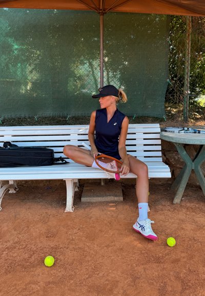 Raqueta de tenis con marco naranja, zapatos blancos y rosas, camiseta sin mangas azul marino y calcetines blancos, sentada en un banco blanco con pelotas de tenis cerca.