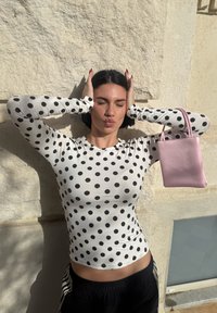 Femme aux yeux fermés tenant ses mains sur la tête, portant une chemise blanche à manches longues avec des pois noirs et portant un petit sac à main rose.