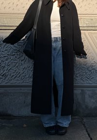 Άτομο που φοράει μακρύ μαύρο παλτό, λευκό τοπ, μπλε τζιν, μαύρα παπούτσια και κρατά μαύρη τσάντα ώμου, στέκεται δίπλα σε υφή τοίχο σε υπαίθριο χώρο.
