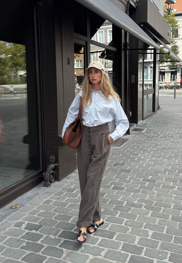 Mujer con cabello largo y rubio que lleva una camisa blanca, pantalones marrones de talle alto, sandalias negras, gorra beige y lleva un bolso marrón caminando por una calle adoquinada.