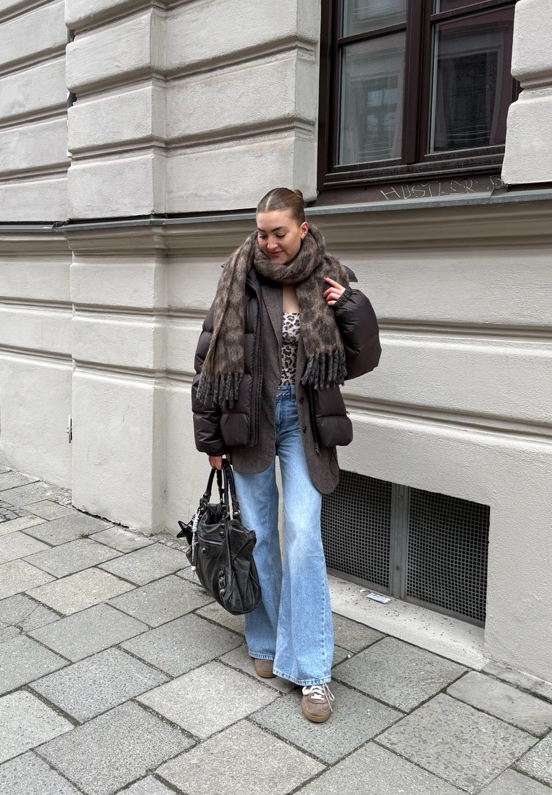 Žena v hnědé prošívané bundě, s topem s leopardím vzorem, velkým šálem, světle modrými džínami a hnědými teniskami, držící černou kabelku na městském chodníku.