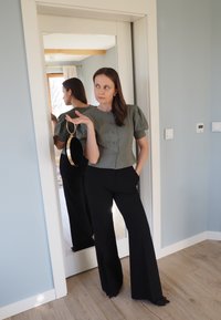 Žalias tekstūruotas trumpas viršus su pūstomis rankovėmis ir sagomis, derinamas su juodomis plačiomis kelnėmis. Laiko apvalią juodą ir aukso spalvos rankinę. Matomas veidrodžio atspindys.