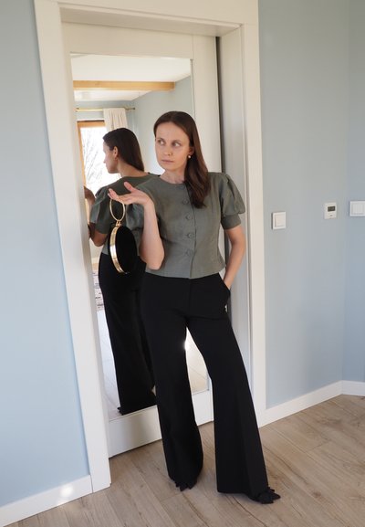 Top recortado verde texturizado con mangas abullonadas y botones, combinado con pantalones negros de pierna ancha. Sosteniendo un bolso redondo negro y dorado. Se observa el reflejo en el espejo.