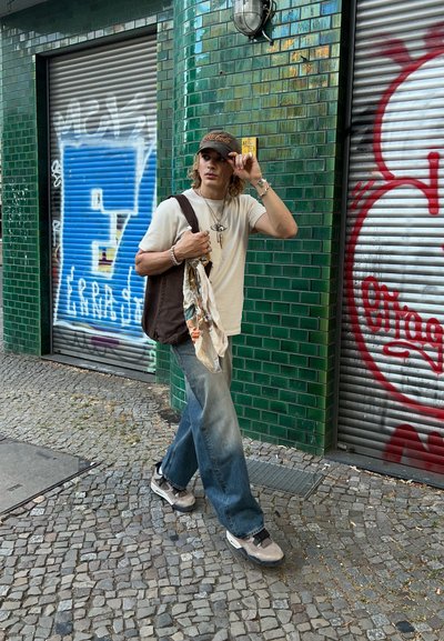 Conjunto casual que incluye una camiseta ligera, vaqueros azules de corte holgado y zapatillas grises. Bolsa de hombro oscura con una bufanda de estampado drapeada sobre ella.