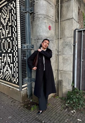 Langer schwarzer Mantel, grauer Pullover und dunkle Hose. Hält eine braune geflochtene Tasche. Steht vor einer strukturierten Steinmauer mit einem gefliesten Muster.