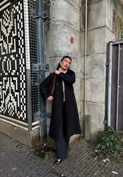 Abrigo negro largo, suéter gris y pantalones oscuros. Sosteniendo un bolso de mimbre marrón. De pie contra una pared de piedra texturizada con un patrón de azulejos.