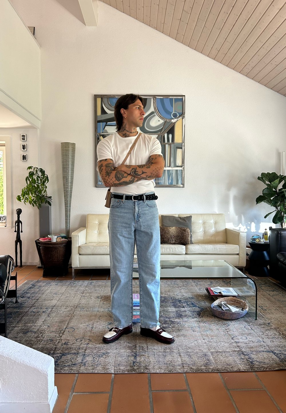 Person i hvit t-skjorte, lyseblå jeans og mørke sko står i et moderne stue med et glasskaffebord og planter.