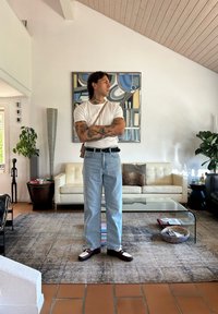 Person i hvit t-skjorte, lyseblå jeans og mørke sko står i et moderne stue med et glasskaffebord og planter.