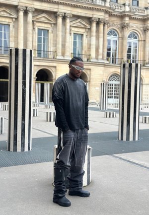 Hombre vestido con ropa negra de gran tamaño y gafas de sol blancas de pie entre columnas altas de rayas blancas y negras en un patio histórico.