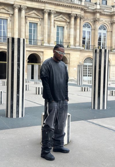 Hombre vestido con ropa negra de gran tamaño y gafas de sol blancas de pie entre columnas altas de rayas blancas y negras en un patio histórico.
