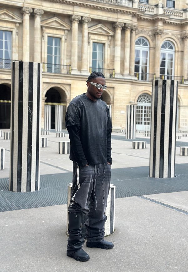 Ein Mann in übergroßer schwarzer Kleidung und weißen Sonnenbrillen steht zwischen hohen schwarz-weiß gestreiften Säulen in einem historischen Innenhof.