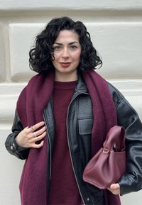 Veste en cuir noire, pull en laine bordeaux et écharpe assortie. Tenant un sac à main texturé bordeaux avec une poignée courte. Cheveux bouclés.