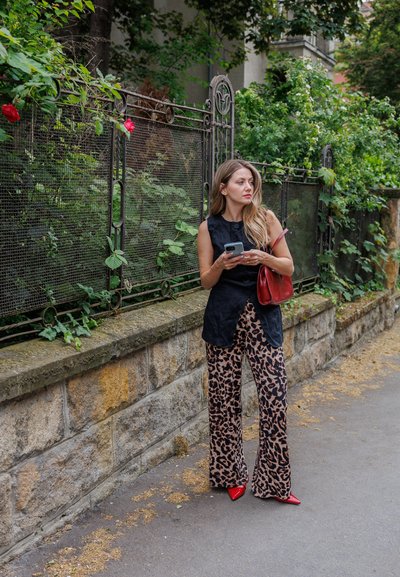Top negro sin mangas, pantalones holgados con estampado de leopardo, tacones rojos de punta y un bolso rojo. El fondo presenta vegetación y un muro de piedra.