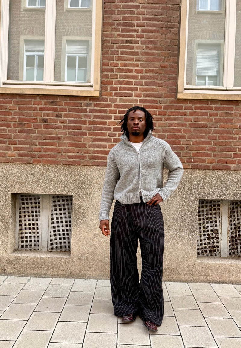 Homme avec des dreadlocks portant un pull gris zippé et un pantalon large noir à fines rayures, debout sur le trottoir contre un mur en briques.
