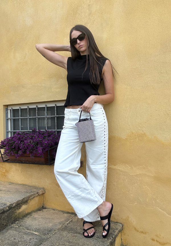 Femme portant un haut sans manches noir et un pantalon blanc à jambes larges, tenant un petit sac à main à carreaux, portant des sandales noires et des lunettes de soleil devant un mur jaune.