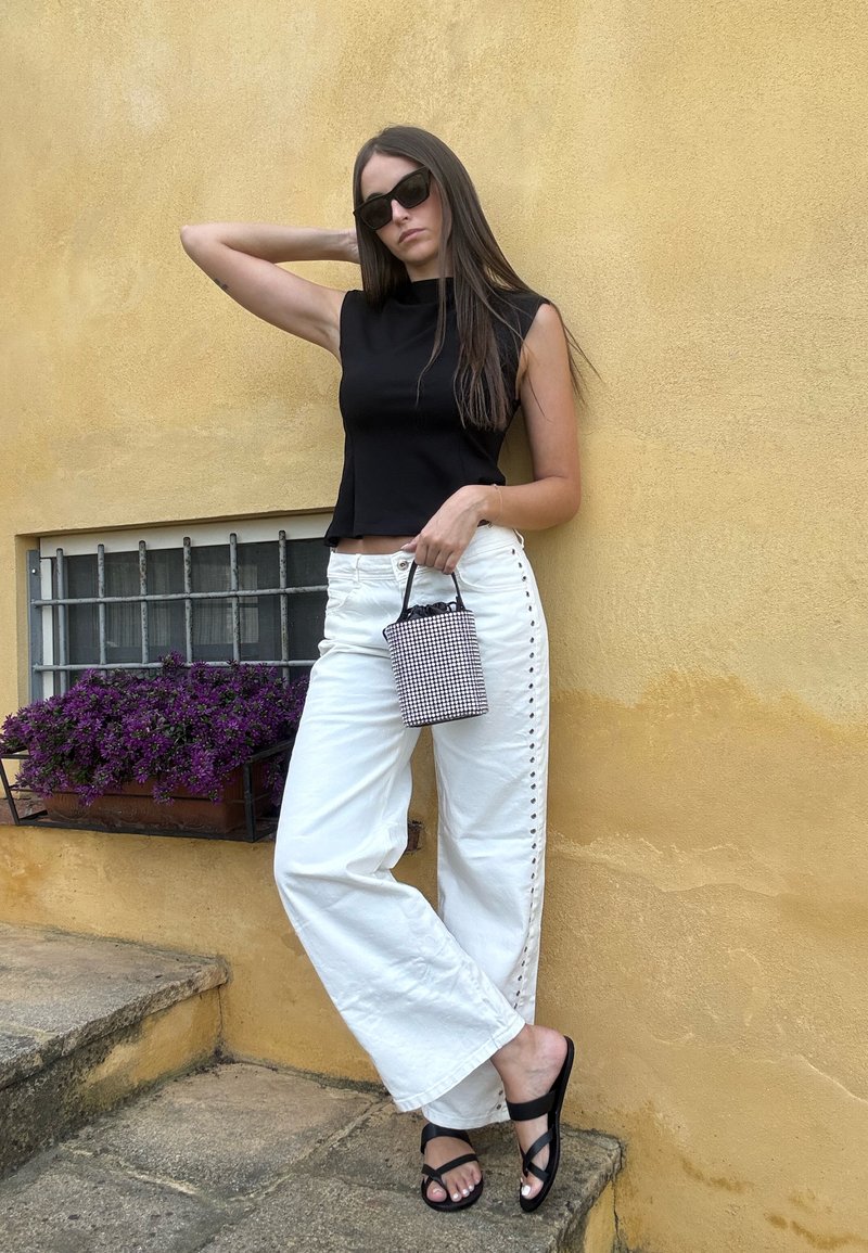 Femme en haut sans manches noir et pantalon blanc à larges jambes tenant un petit sac à main à carreaux, portant des sandales noires et des lunettes de soleil devant un mur jaune.
