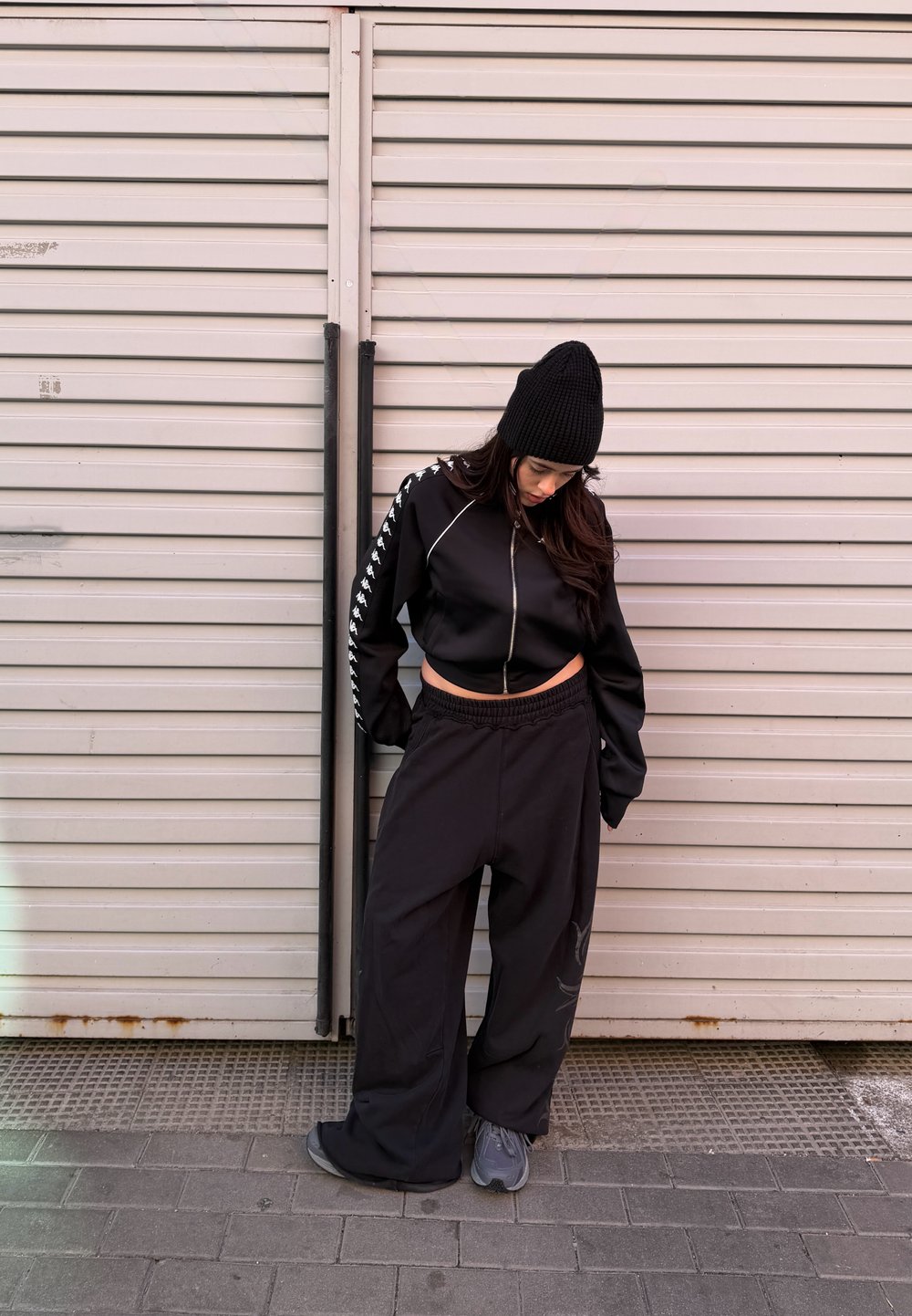 Femme portant un bonnet noir, une veste zippée courte et un pantalon large noir, regardant vers le bas devant une porte métallique rose clair à volets.