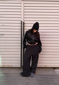 Femme portant un bonnet noir, une veste zippée courte et un pantalon large noir, regardant vers le bas devant une porte métallique rose clair à volets.