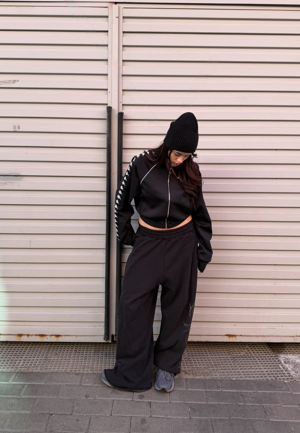 Mustas mütsis naine, lühike lukuga jope ja laiad mustad püksid, seisab ja vaatab alla heleroosa metallist ruloovärava taustal.