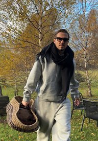 Felpa grigia in pile e pantaloni della tuta indossati da una persona che porta un cesto intrecciato con strisce scure, sullo sfondo di fogliame autunnale e sedie.