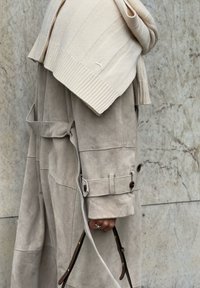 Trench-coat en daim beige avec une taille ceinturée et des poignets boutonnés, superposé avec un pull en maille côtelée crème drapé sur les épaules.