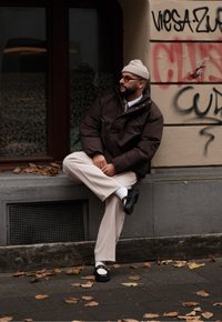 Καφέ μπουφάν, ανοιχτό μπλε πουκάμισο, μπεζ παντελόνι, μαύρα παπούτσια με λευκές λεπτομέρειες. Καθισμένος σε ένα βαθούλωμα, περιτριγυρισμένος από ξεραμένα φύλλα και γκράφιτι.