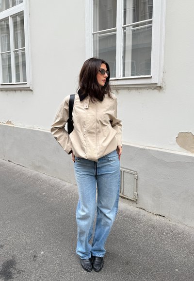 Chaqueta beige con cuello negro, de corte holgado, combinada con vaqueros azul claro de pierna ancha y zapatos negros. De pie frente a una pared de color claro.