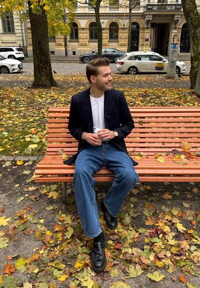 Joven con un blazer negro a rayas y camisa blanca, sentado en un banco de madera, sosteniendo una taza, rodeado de hojas de otoño.