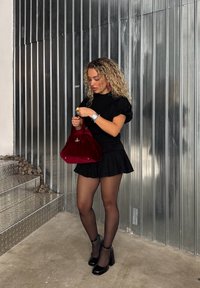 Svart topp med puffede ermer, plisserte mini-skirt, gjennomsiktige strømpebukser og svarte h�æler. Holder en rød velvettveske med gullfester. Metallisk bakgrunn.