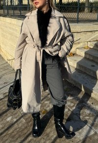 Beige trench coat med krage i svart syntetpäls, svarta sidostråk, grå byxor och glänsande svarta platåstövlar, håller en svart handväska.