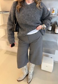 Γυναίκα που φοράει γκρίζο πουλόβερ με υφή, φαρδιά γκρίζα παντελόνια, λευκά καουμπόικα μποτάκια με μοτίβο φιδιού, ασημένια βραχιόλια και κρατάει μια μαύρη χειραποσκευή.