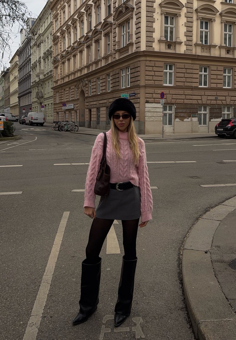 Femme portant un pull rose en maille torsadée, une mini-jupe noire, des collants, de longues bottes noires, un chapeau noir et des lunettes de soleil, debout à un coin de rue en ville.