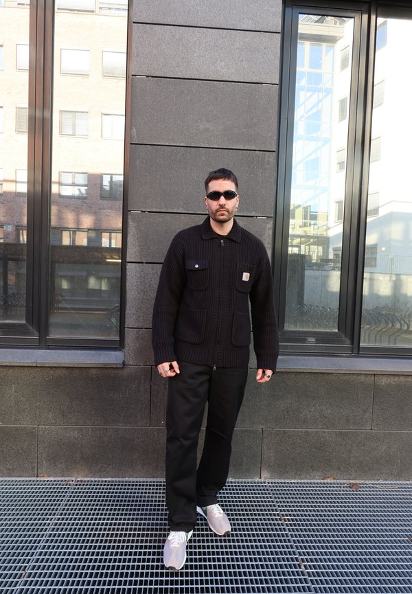 Homme portant un pull noir, un pantalon noir, des baskets blanches et des lunettes de soleil, debout contre un mur carrelé sombre avec des fenêtres reflétant des bâtiments urbains.