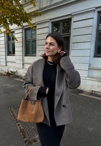 Hnedá semišová kabelka, šedý vlnený blazer s širokými klopami, čierne pletené šaty. Exteriérové prostredie s budovou v pozadí.