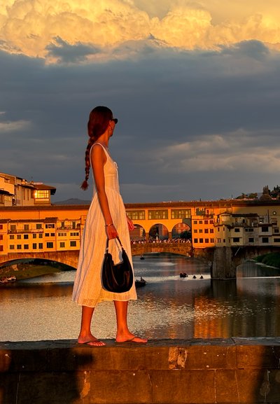 Vestido blanco texturizado que llega a la mitad de la pantorrilla; cabello trenzado; bolso negro; de pie al lado de un río al atardecer; puente histórico al fondo.