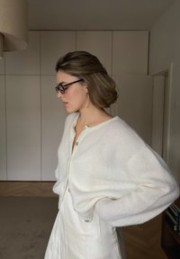 Biały, dzianinowy kardigan z guziczkami z przodu, o luźnym kroju i miękkiej fakturze, noszony na kremowej sukience, w jasnym wnętrzu.