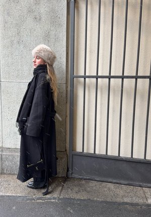 Mujer con abrigo negro y bufanda, con gorro de piel, de pie junto a un muro de piedra y una puerta metálica sobre un pavimento mojado.