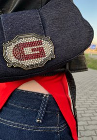 Denim crossover-veske med mørkeblå ytre, prydet med et rhinestone "G"-logo i rødt og sølv, med en strukturert design og dekorativt hardware.