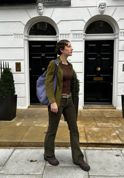 Chaqueta de ante verde oliva, blusa marrón ajustada, pantalones de pana verde oscuro acampanados, bolso de mano con patrón púrpura y botas de plataforma marrones.