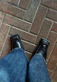 Chaussures noires en cuir à enfiler avec des bouts carrés, des coutures détaillées et des semelles épaisses, portées avec un jean en denim bleu, posées sur un pavé en briques texturé.