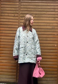 Blommig quiltad jacka med vinröd krage och mudd, kombinerad med en lång mörk klänning och en rosa handväska. Träbakgrund.