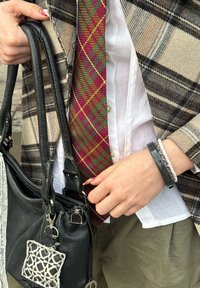 Άτομο με πουκάμισο σε λευκό χρώμα και σακάκι καρό προσαρμόζει γραβάτα με κόκκινο και πράσινο μοτίβο, κρατώντας μαύρη δερμάτινη τσάντα με μενταγιόν με σχέδιο.