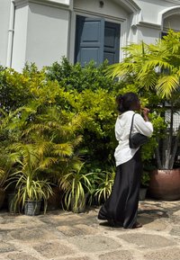 Kvinne med svart skjørt, hvit skjorte og svart veske som går forbi potteplanter i fronten av en hvit bygning med mørk dør.