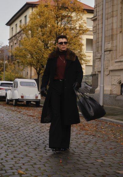 Abrigo negro con cuello de piel, suéter de cuello alto color granate, pantalones negros y guantes; lleva un gran bolso negro de mano, caminando por una calle empedrada.