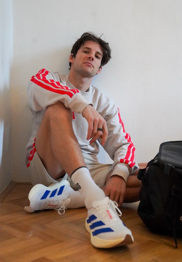 Jeune homme en tenue de sport blanche et rouge assis sur un sol en bois contre un mur blanc, avec un sac à dos noir à proximité.