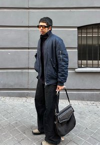 Άνδρας με σκοτεινά γυαλιά ηλίου, μπλε μπουφάν bomber, μαύρο παντελόνι, κρατώντας μια μεγάλη μαύρη τσάντα χε ιρός, στέκεται σε γκρι πεζοδρόμιο.