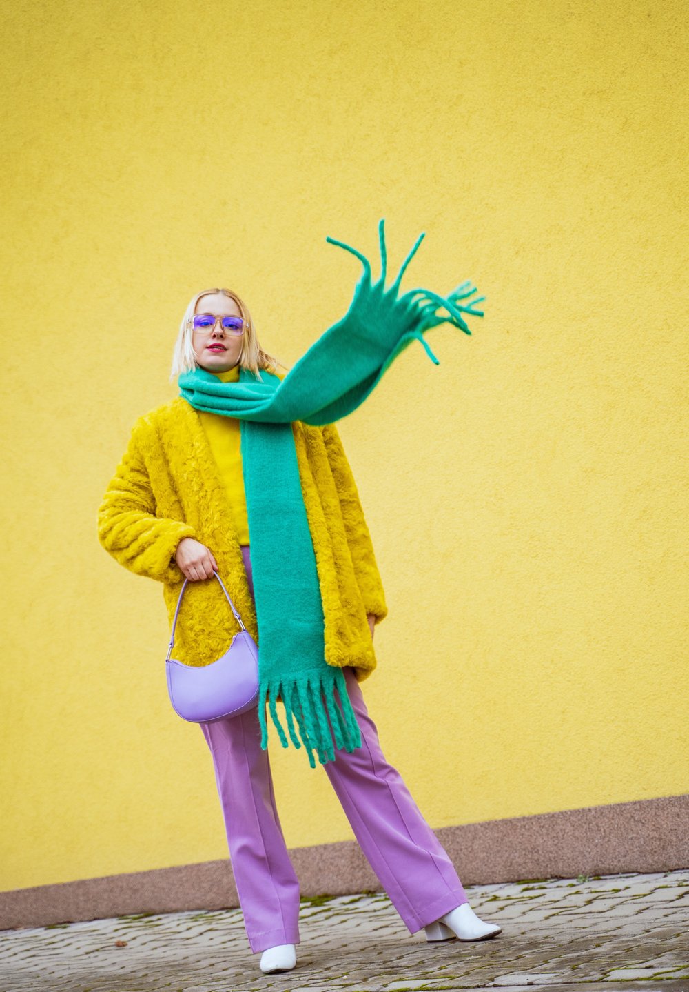 Žlutý umělý kožich, živě zelený pletený šátek, fialová blůza a kalhoty, levandulová taška, bílé boty, modelka před žlutou zdí.