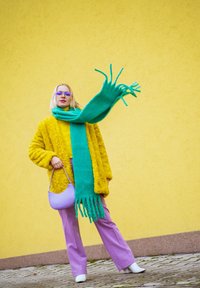 Žlutý umělý kožich, živě zelený pletený šátek, fialová blůza a kalhoty, levandulová taška, bílé boty, modelka před žlutou zdí.