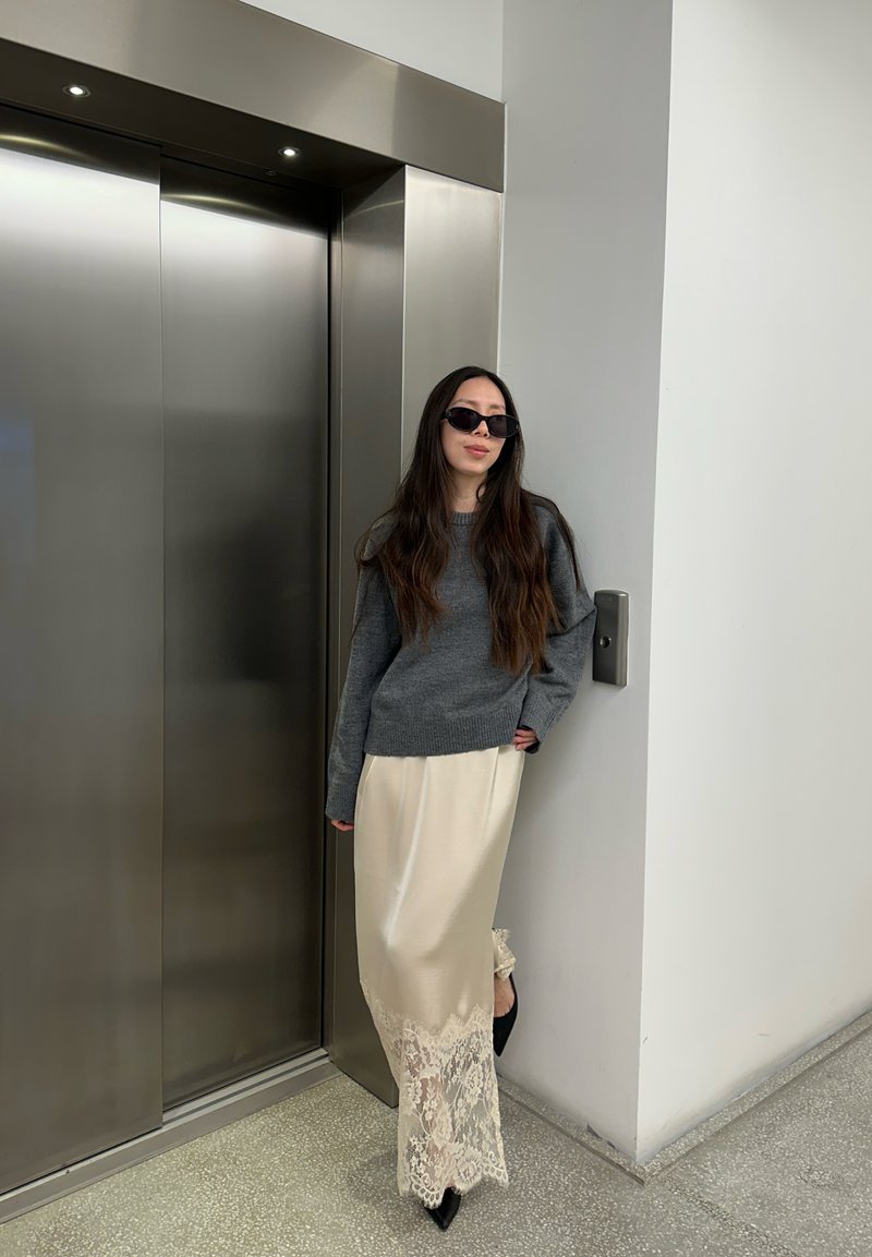 Donna con capelli lunghi che indossa occhiali da sole, maglione grigio e gonna color crema con bordi in pizzo, in piedi accanto a porte chiuse di ascensore in acciaio inox.