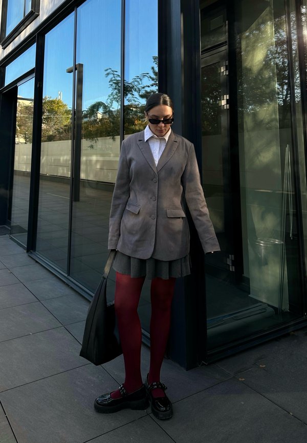 Szary żakiet na białej koszuli, zestawiony ze plisowaną szara spódnicą. Bordowe rajstopy i czarne masywne buty, trzymająca czarną torbę. Szklane okna w tle.